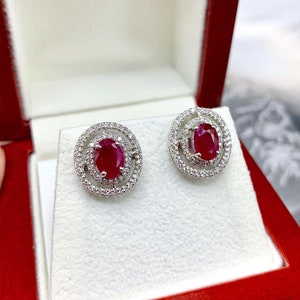 May include: A pair of silver earrings with a red oval ruby gemstone surrounded by a halo of smaller white gemstones.