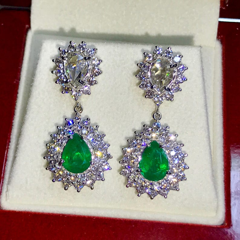 May include: A pair of emerald and diamond drop earrings. The earrings feature a pear-shaped emerald surrounded by a halo of round diamonds. The emeralds are a vibrant green color and the diamonds are sparkling white.
