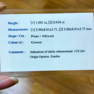 May include: A white card with black text describing a green pear-shaped gemstone. The card lists the weight as 1.001 carats and 0.934 carats, the measurements as 5.99 x 8.01 x 3.71 mm and 5.80 x 8.01 x 3.77 mm, the shape as pear and the cut as mixed. The card also notes that the gemstone has indications of clarity enhancement and that the origin is Zambia.