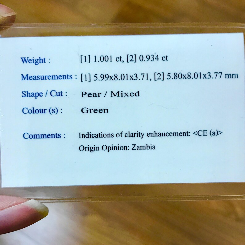 May include: A white card with black text describing a green pear-shaped gemstone. The card lists the weight as 1.001 carats and 0.934 carats, the measurements as 5.99 x 8.01 x 3.71 mm and 5.80 x 8.01 x 3.77 mm, the shape as pear and the cut as mixed. The card also notes that the gemstone has indications of clarity enhancement and that the origin is Zambia.