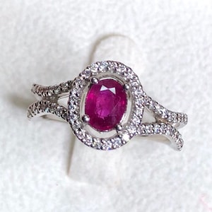 May include: A silver ring featuring a large, oval-cut ruby gemstone surrounded by a halo of small, clear gemstones. The ring's band splits into two delicate, curving arms on either side of the halo, creating a unique design. The ruby is a deep, rich red color.