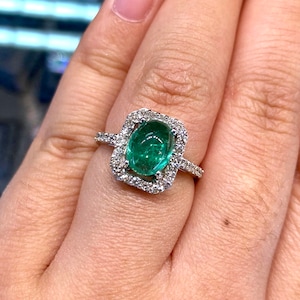 Könnte beinhalten: Ein Ring mit einem großen, ovalen Smaragd, der in einen quadratischen Rahmen aus kleineren Diamanten gefasst ist. Der Ring wird an einem Finger getragen.