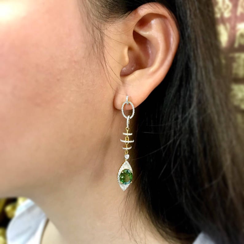 May include: Gold dangle earrings with a green gemstone and diamond accents. The earrings feature a chain-like design with multiple small circles.