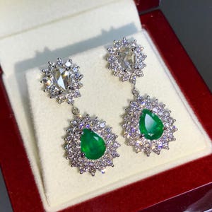 May include: A pair of dangle earrings with pear-shaped emerald gemstones surrounded by a halo of smaller diamonds. The earrings are suspended from a smaller pear-shaped diamond surrounded by a halo of smaller diamonds.