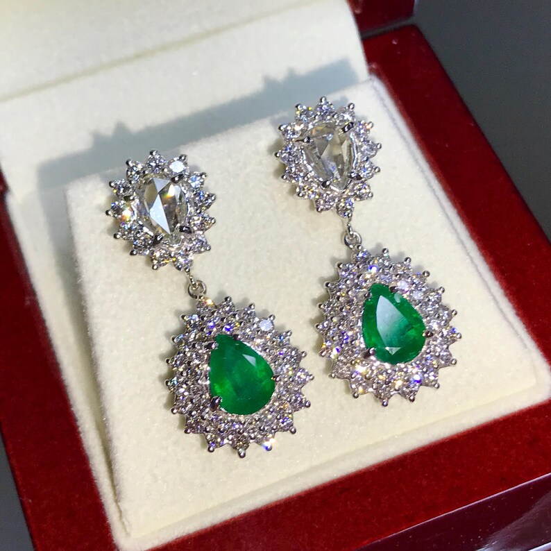 May include: A pair of dangle earrings with pear-shaped emerald gemstones surrounded by a halo of smaller diamonds. The earrings are suspended from a smaller pear-shaped diamond surrounded by a halo of smaller diamonds.