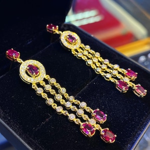 May include: A pair of gold earrings with dangling ruby and diamond accents. The earrings feature a round halo design with a ruby center stone and a row of diamonds surrounding it. The ruby and diamond accents dangle from the halo design.