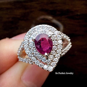 May include: A pear-shaped ruby ring set in silver with a double halo of smaller diamonds. The ring is on a finger.