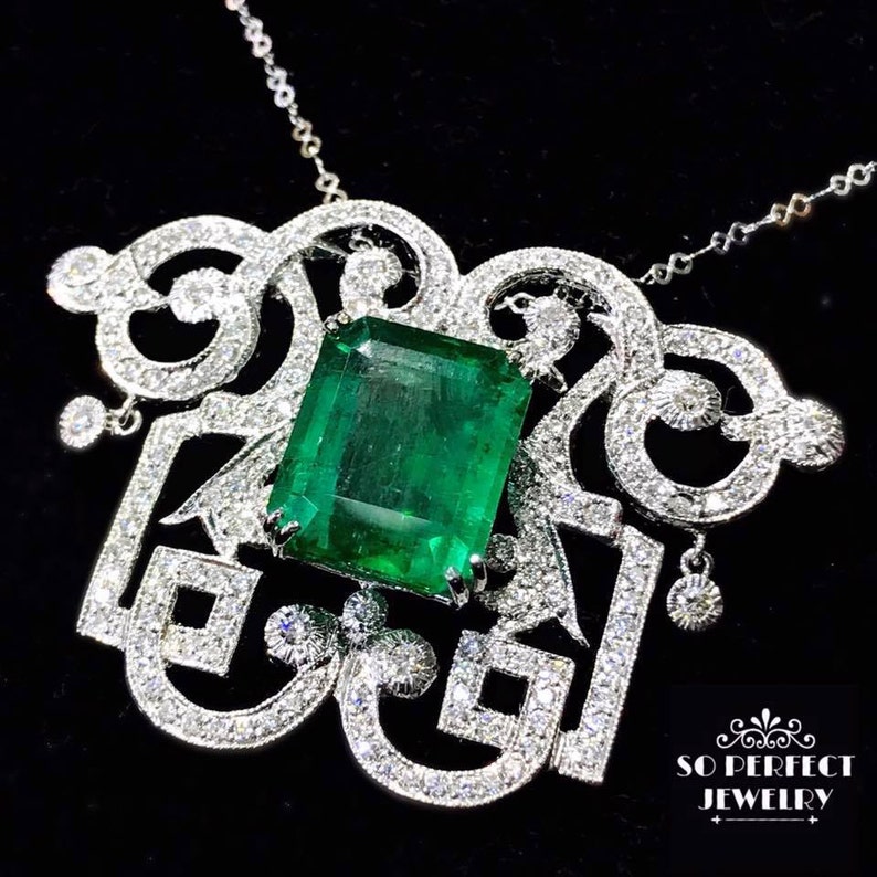 May include: A silver pendant necklace with a large emerald green gemstone set in a intricate, diamond-encrusted design. The pendant features a delicate, ornate pattern with a dangling diamond accent.