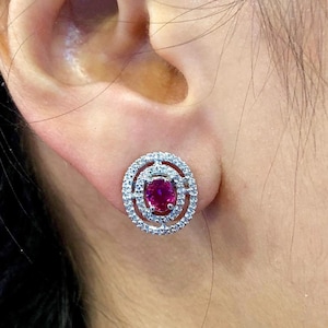 May include: A pair of silver earrings with a red oval gemstone surrounded by a halo of smaller white gemstones.