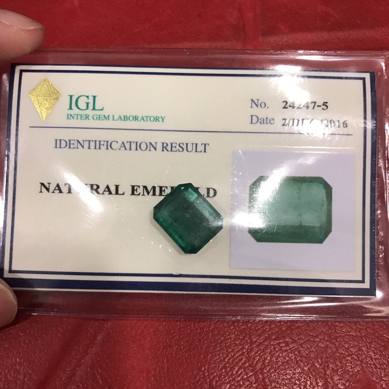 May include: A green emerald gemstone with a rectangular shape, certified by the IGL Inter Gem Laboratory. The certificate shows the identification result as "NATURAL EMERALD" with the number 24247-5 and date 2/11/2016.