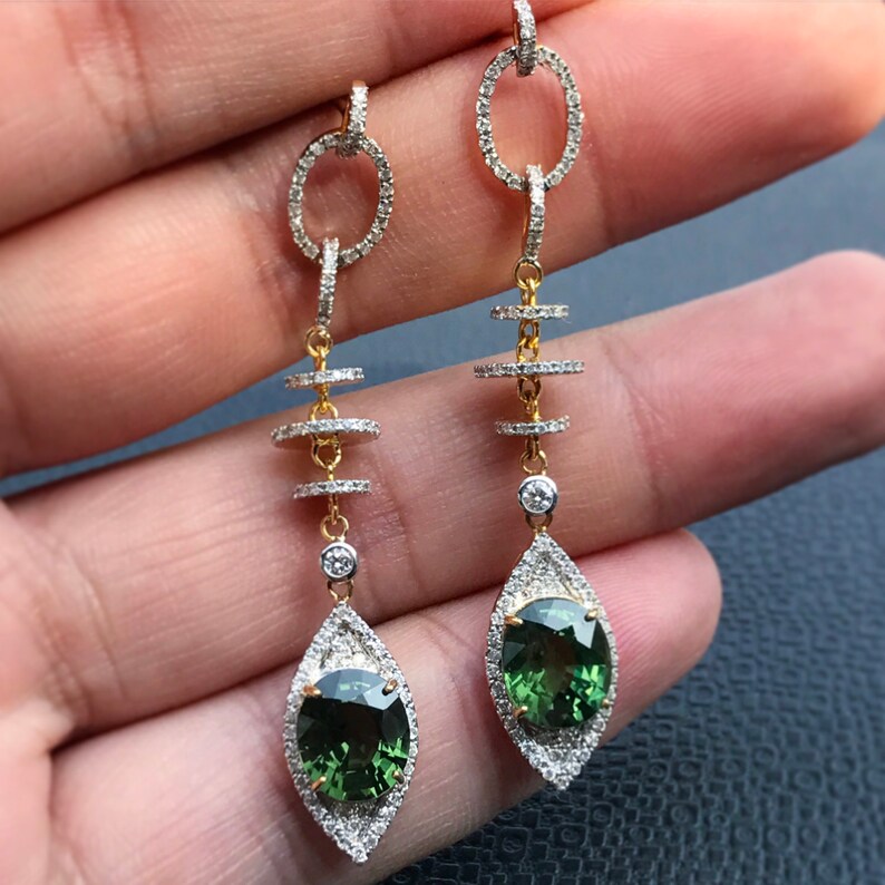 May include: A pair of long, dangling earrings with green gemstones and diamond accents. The earrings feature a chain with three circular links and a teardrop-shaped pendant with a green gemstone.