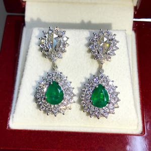 May include: A pair of dangle earrings with pear-shaped emerald gemstones surrounded by a halo of smaller diamonds. The earrings are suspended from a smaller pear-shaped diamond surrounded by a halo of smaller diamonds.