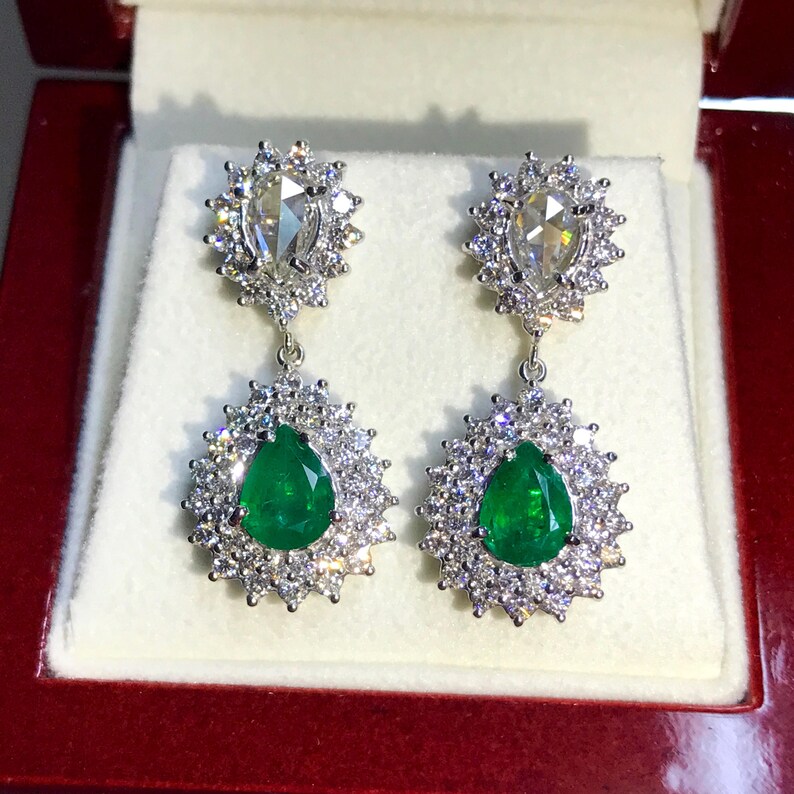 May include: A pair of dangle earrings with pear-shaped emerald gemstones surrounded by a halo of smaller diamonds. The earrings are suspended from a smaller pear-shaped diamond surrounded by a halo of smaller diamonds.