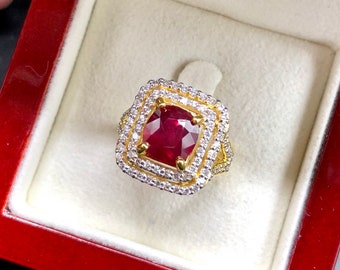May include: A gold ring with a large, square-cut ruby center stone. The ruby is surrounded by two layers of smaller, round, white gemstones. The ring is set in a red jewelry box with a cream-colored interior.