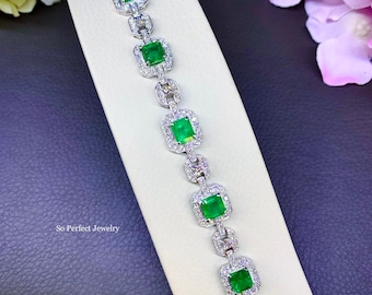 May include: A white gold bracelet with a row of emerald green square-shaped gemstones set in a diamond halo. The bracelet is on a white display stand. "So Perfect Jewelry" is printed on the stand.
