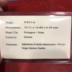 May include: A green gemstone with a weight of 9.813 carats and measurements of 12.11 x 14.60 x 6.58 millimeters. The gemstone is an octagon step cut. The label indicates that the gemstone has been clarity enhanced and is of Zambian origin.