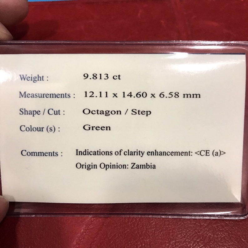 May include: A green gemstone with a weight of 9.813 carats and measurements of 12.11 x 14.60 x 6.58 millimeters. The gemstone is an octagon step cut. The label indicates that the gemstone has been clarity enhanced and is of Zambian origin.
