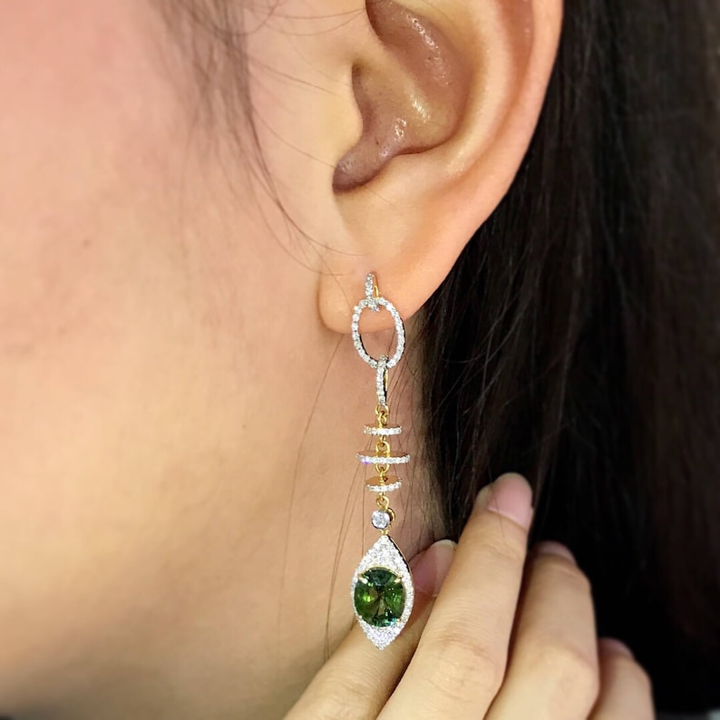 May include: A pair of dangle earrings with a green gemstone and diamond accents. The earrings feature a gold chain with a circular design and a small diamond at the bottom.