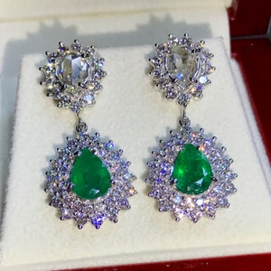 May include: A pair of dangle earrings with pear-shaped emerald gemstones surrounded by a halo of smaller diamonds. The top of each earring features a round, rose-cut diamond surrounded by a halo of smaller diamonds.