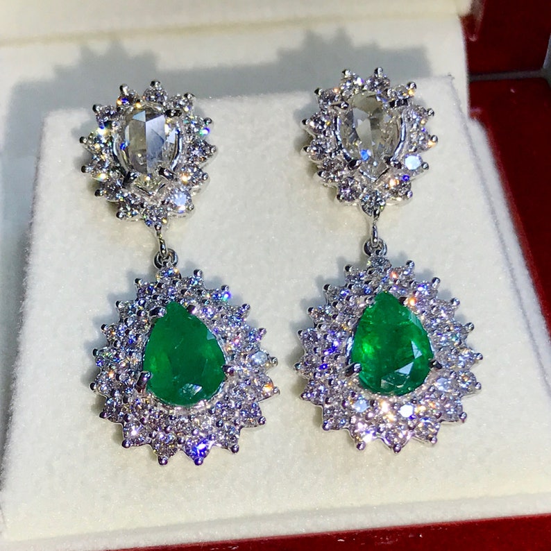May include: A pair of dangle earrings with pear-shaped emerald gemstones surrounded by a halo of smaller diamonds. The top of each earring features a round, rose-cut diamond surrounded by a halo of smaller diamonds.