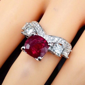 以下が含まれることがあります： 大きなクッションカットの赤いルビーを、小さな白いダイヤモンドで囲んだリング。リングはホワイトゴールド製で、繊細で複雑なデザインです。