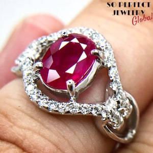 May include: A silver ring with a large oval ruby gemstone surrounded by smaller diamonds. The ring is set in a delicate, openwork design.