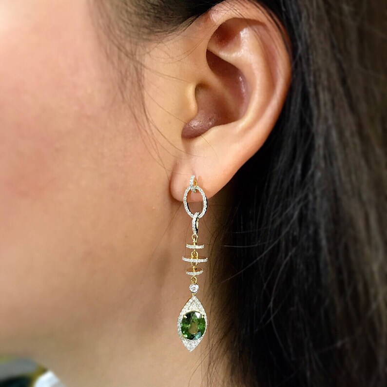 May include: A pair of dangle earrings with a green gemstone and diamond accents. The earrings feature a chain link design with a geometric shape and a teardrop-shaped pendant.