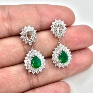 May include: A pair of pear-shaped emerald and diamond dangle earrings. The earrings feature a large pear-shaped emerald surrounded by a halo of smaller diamonds. The emeralds are a vibrant green color and the diamonds are clear and sparkly.