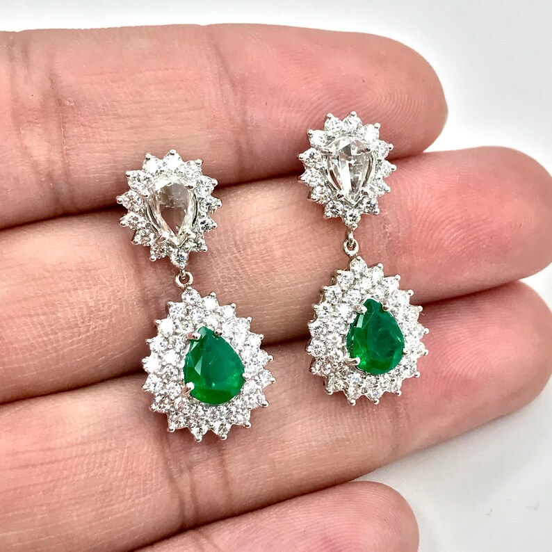May include: A pair of pear-shaped emerald and diamond dangle earrings. The earrings feature a large pear-shaped emerald surrounded by a halo of smaller diamonds. The emeralds are a vibrant green color and the diamonds are clear and sparkly.