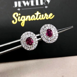 May include: A pair of oval ruby earrings with a halo of small, clear gemstones. The earrings are held by a silver tweezer against a black background. The word "Signature" is written in yellow cursive.