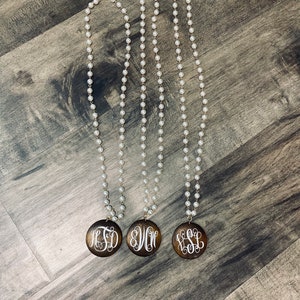 May include: Three long pearl necklaces with wooden pendants. Each pendant features a different white monogram against a dark brown background. The necklaces are displayed on a wooden surface.
