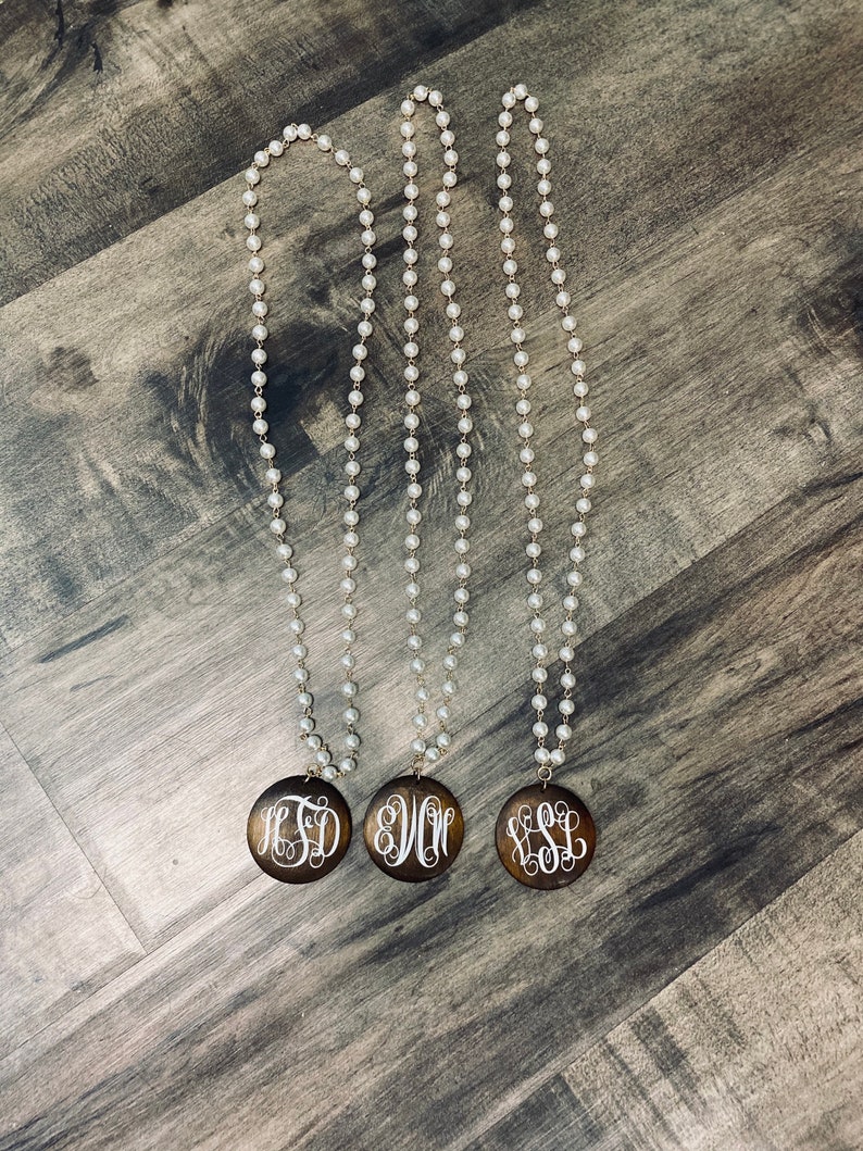 May include: Three long pearl necklaces with wooden pendants. Each pendant features a different white monogram against a dark brown background. The necklaces are displayed on a wooden surface.