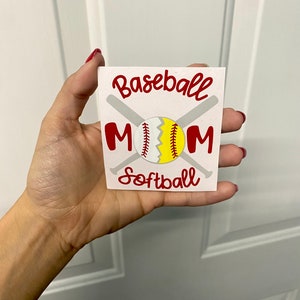 May include: A white square with the words "Baseball Mom Softball" in red script. The design includes a baseball and softball, with crossed bats in the background. The baseball is split, showing a yellow and white design.