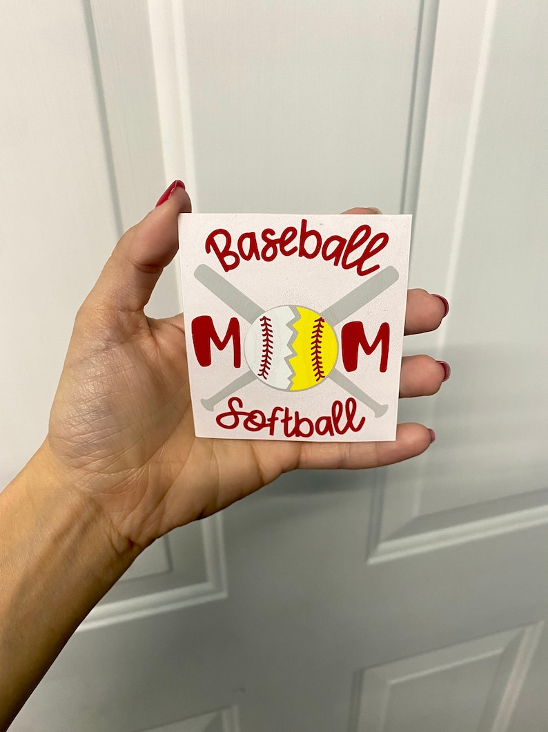 May include: A white square with the words "Baseball Mom Softball" in red script. The design includes a baseball and softball, with crossed bats in the background. The baseball is split, showing a yellow and white design.