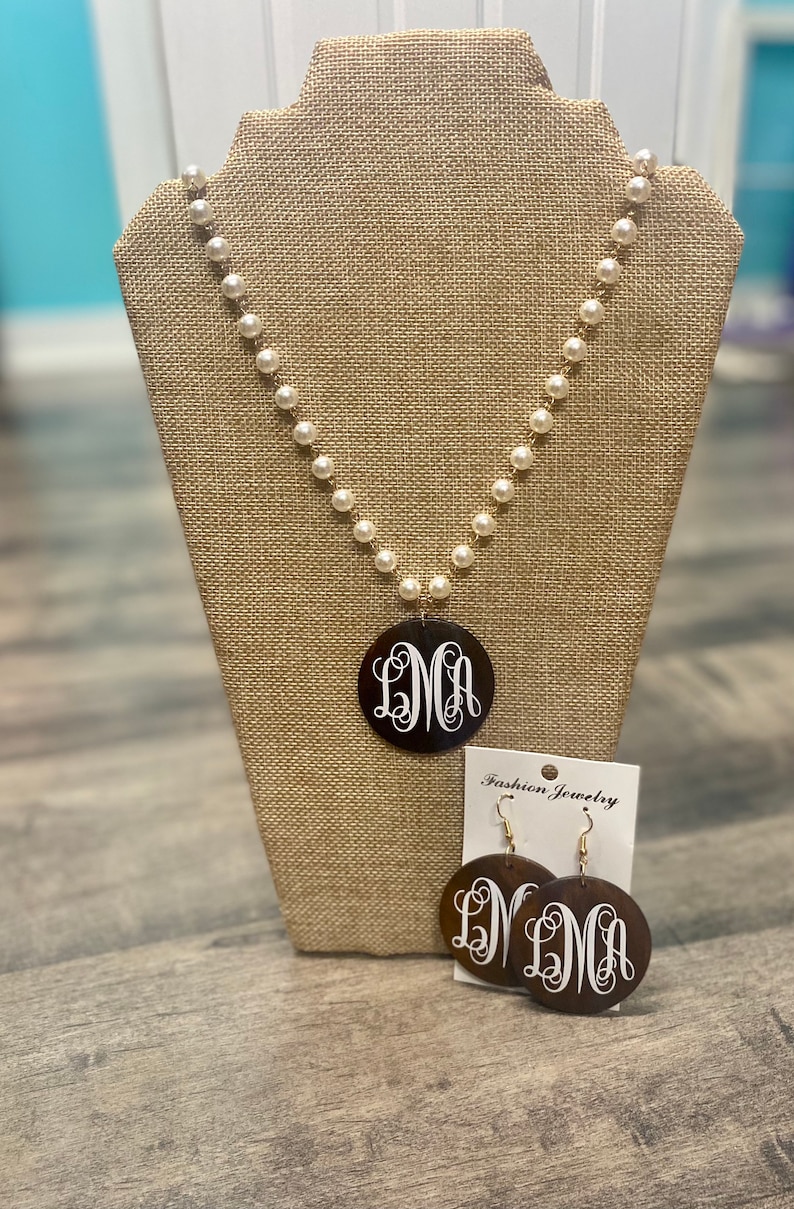 May include: A pearl necklace with a gold chain and a large, round, dark brown pendant featuring the white monogram "LMA". Matching earrings with the same monogram are displayed on a Fashion Jewelry card.