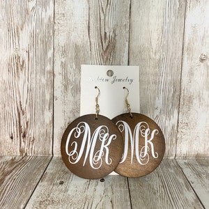 May include: Wooden round earrings with a dark brown finish. Each earring features a white monogram of the letters "MK" in a cursive font. The earrings hang from gold-colored fish hook ear wires. The earrings are displayed on a white card that reads "Fashion Jewelry".