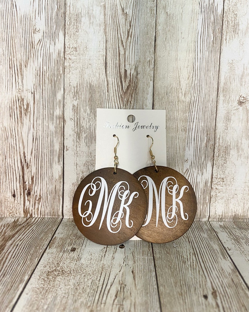 May include: Wooden round earrings with a dark brown finish. Each earring features a white monogram of the letters "MK" in a cursive font. The earrings hang from gold-colored fish hook ear wires. The earrings are displayed on a white card that reads "Fashion Jewelry".