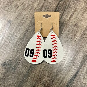May include: White baseball-themed teardrop earrings with red stitching and the number 09 in black. The earrings are displayed on a brown cardboard earring card with silver hooks.