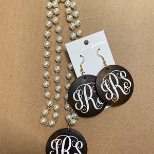 May include: A jewelry set featuring a pearl necklace and matching earrings. The earrings and pendant are dark brown wood with a white monogram. The earrings hang from gold-colored hooks. The necklace has a gold chain.