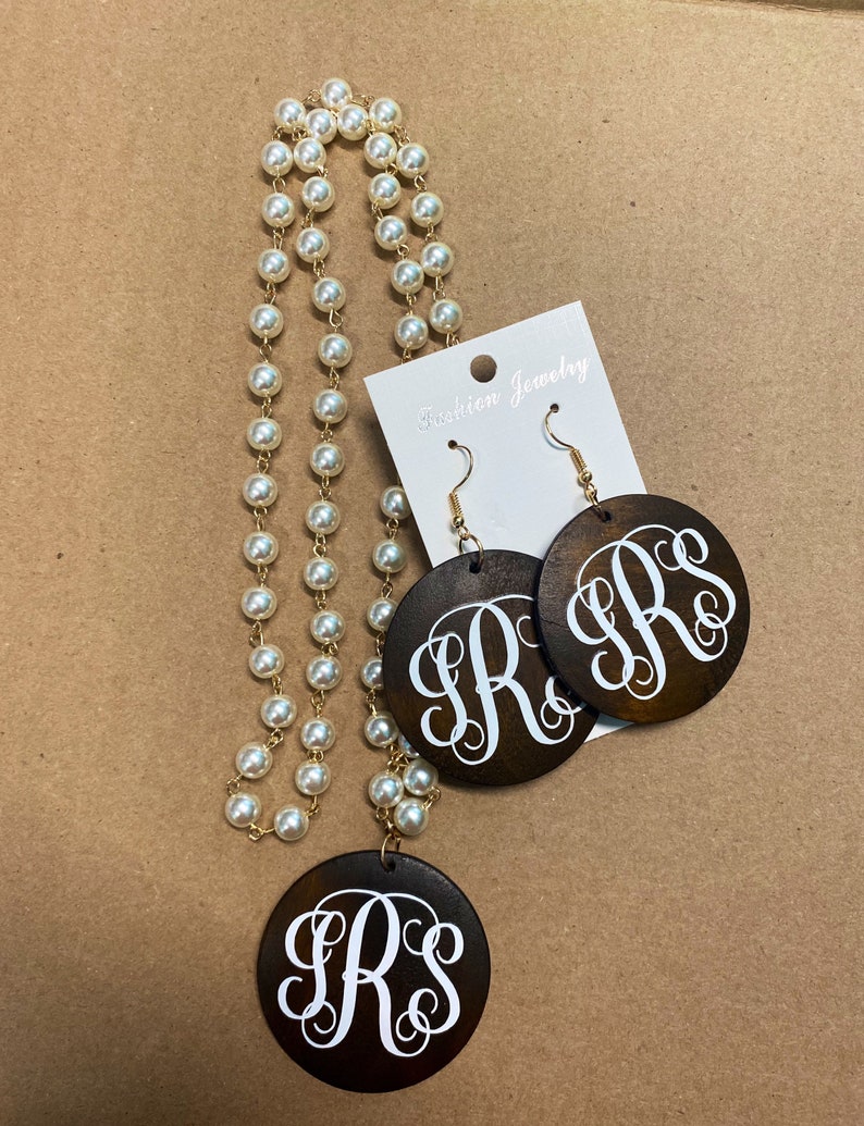 May include: A jewelry set featuring a pearl necklace and matching earrings. The earrings and pendant are dark brown wood with a white monogram. The earrings hang from gold-colored hooks. The necklace has a gold chain.