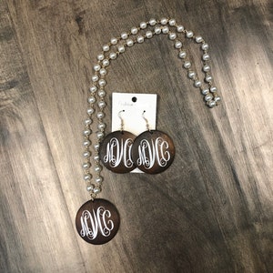 May include: A necklace and earring set featuring a pearl strand with a gold-tone chain. The pendant and earrings are dark brown wooden circles with a white monogram design. The earrings are hanging from silver-tone hooks.