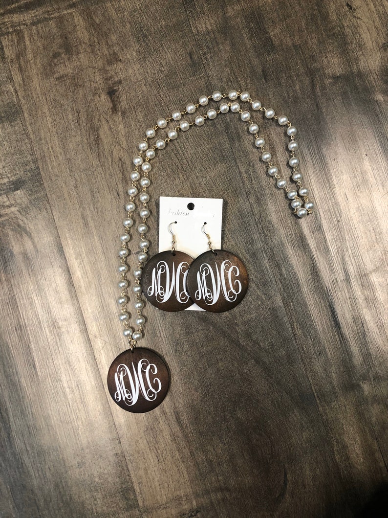 May include: A necklace and earring set featuring a pearl strand with a gold-tone chain. The pendant and earrings are dark brown wooden circles with a white monogram design. The earrings are hanging from silver-tone hooks.