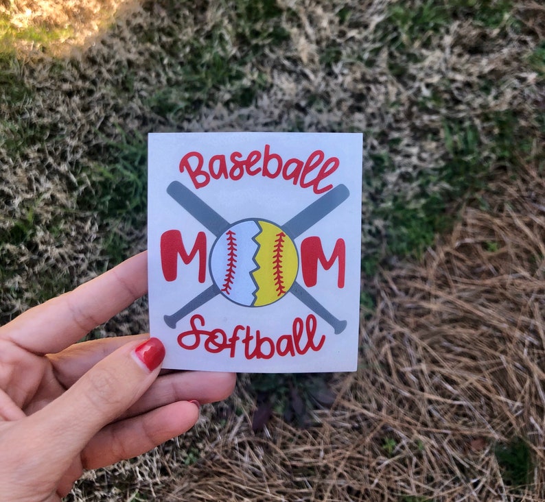 May include: A white square sticker with the words "Baseball Mom Softball" in red script. The word "MOM" is in large red letters. A baseball and two crossed bats are in the center of the sticker.