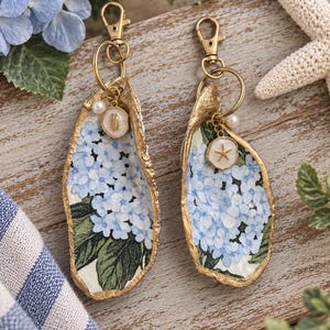 Può includere: Due portachiavi decorativi a forma di conchiglia di ostrica con un motivo floreale di ortensie blu all'interno e bordi dipinti in oro. Ogni conchiglia include un moschettone dorato, una piccola perla bianca e un piccolo ciondolo.