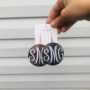 May include: Dark brown wooden earrings with a white monogram design. The earrings are round with gold-colored hooks. The earrings are held up by a white card that says "Fashion Jewelry".