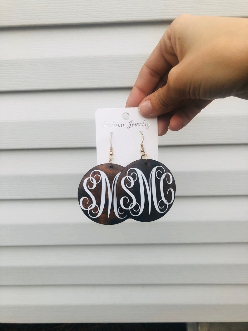 May include: Dark brown wooden earrings with a white monogram design. The earrings are round with gold-colored hooks. The earrings are held up by a white card that says "Fashion Jewelry".