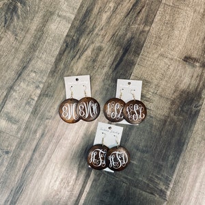May include: Three pairs of wooden monogram earrings. Each pair features a dark brown, circular wooden base with a white, cursive monogram design. The earrings are displayed on white earring cards with the text "Fashion Jewelry".