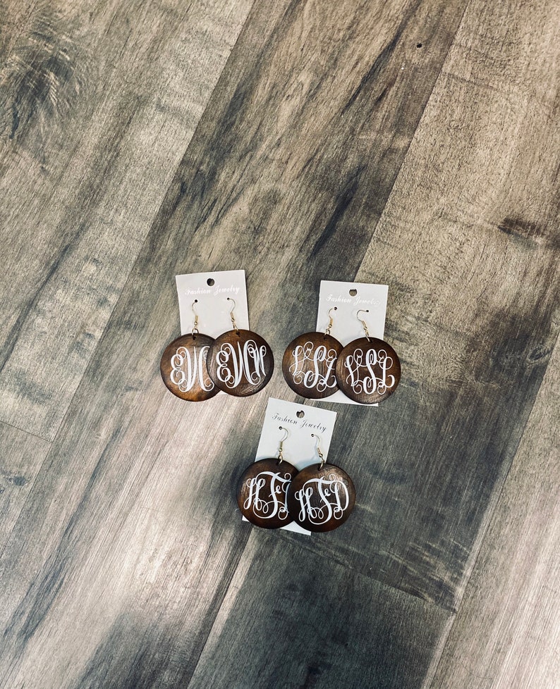 May include: Three pairs of wooden monogram earrings. Each pair features a dark brown, circular wooden base with a white, cursive monogram design. The earrings are displayed on white earring cards with the text "Fashion Jewelry".