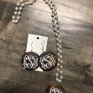 May include: A jewelry set featuring a long pearl necklace with a gold chain, matching earrings, and a pendant. The earrings and pendant are dark brown, circular, and feature a white monogram. The jewelry is displayed on a wooden surface.