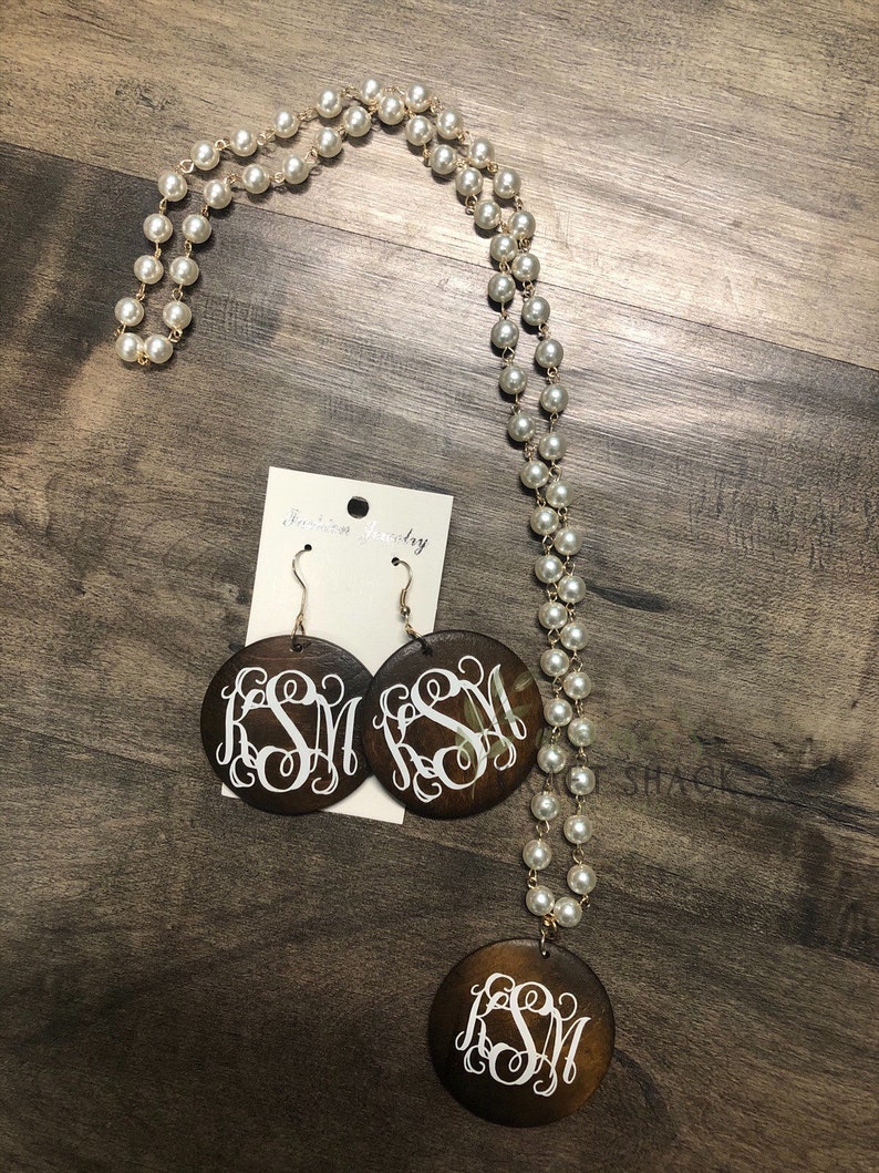 May include: A jewelry set featuring a long pearl necklace with a gold chain, matching earrings, and a pendant. The earrings and pendant are dark brown, circular, and feature a white monogram. The jewelry is displayed on a wooden surface.